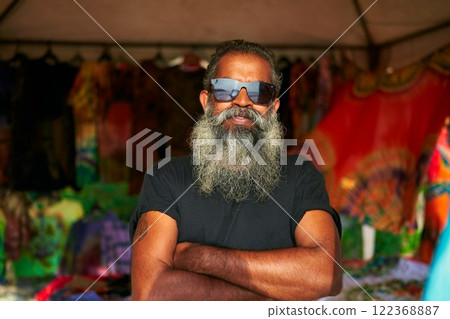 Senior artist with big beard, sunglasses stands in front of colorful tie-dye t-shirts at vibrant fair. Bearded man crosses arms, smiles confidently. Artisan spirit in open-air market. Senior artist with big beard, sunglasses stands in front of colorful tie-dye t-shirts at vibrant fair. Bearded man crosses arms, smiles confidently. Artisan spirit in open-air market. 122368887