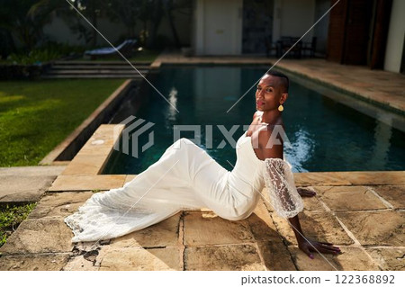 Gender fluid person in wedding dress relaxes by pool at villa. Elegant bride lounges on stone edge. Celebrate love in modern wedding. Luxury villa setting for LGBTQ ceremony. Beautiful bridal fashion. 122368892