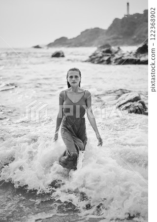 Woman in wet dress walks through ocean waves on rocky beach. Confident female embodies femininity and beauty with serene expression. Natural scenery suggests travel, adventure, summer vibes. 122368902