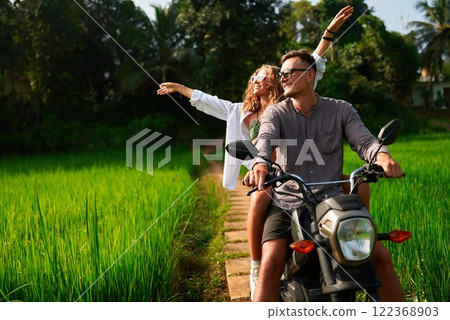 Couple rides motorbike through lush rice fields. Enthusiastic woman spreads arms. Man drives scooter on narrow path. Vibrant green landscape, sunny day. Safe journey exploring nature. 122368903