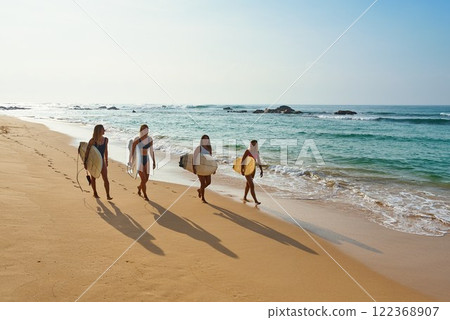Four friends walk with surfboards on sandy beach. Young surfers enjoy sunny ocean day. Group heads to sea waves for surfing adventure. Coastal travel, warm weather, active lifestyle. Four friends walk with surfboards on sandy beach. Young surfers enjoy sunny ocean day. Group heads to sea waves for surfing adventure. Coastal travel, warm weather, active lifestyle. 122368907
