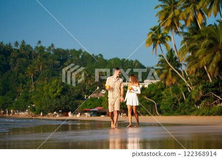 Couple holds hands on sandy beach at sunset, sipping coconut water. Romantic walk along sea shore, man and woman in summer clothes enjoy tropical vacation. Love, travel, leisure on exotic beach. 122368914