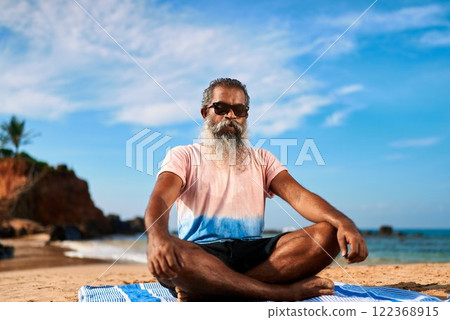 Elderly man with sunglasses meditates in lotus pose on beach mat. Stylish senior doing yoga outdoors at ocean shore. Wellness, health, relaxation for active lifestyle. Summer sky, sea view. 122368915