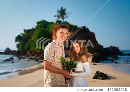 Romantic couple stands on tropical beach at sunset, man holds bouquet, embracing woman. Smiling lovers enjoy Valentines Day, posing for memory. Happiness, love, travel concept. Romantic couple stands on tropical beach at sunset, man holds bouquet, embracing woman. Smiling lovers enjoy Valentines Day, posing for memory. Happiness, love, travel concept. 122368918