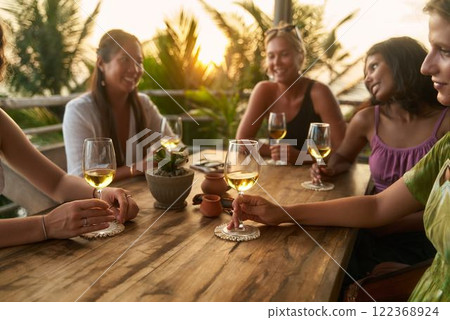 Women enjoy evening wine in tropical restaurant. Friends laugh, share stories, celebrate bachelorette party traveling. Relaxed meeting of diverse women. Sunset scenery adds warm glow. Women enjoy evening wine in tropical restaurant. Friends laugh, share stories, celebrate bachelorette party traveling. Relaxed meeting of diverse women. Sunset scenery adds warm glow. 122368924