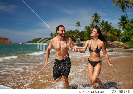 Joyful couple runs along tropical beach, hand in hand, splashing water. Shirtless man, bikini-clad woman enjoy seaside run, laughing. Fitness holiday fun blend in sunny adventure, wellness, escape. Joyful couple runs along tropical beach, hand in hand, splashing water. Shirtless man, bikini-clad woman enjoy seaside run, laughing. Fitness holiday fun blend in sunny adventure, wellness, escape. 122368926