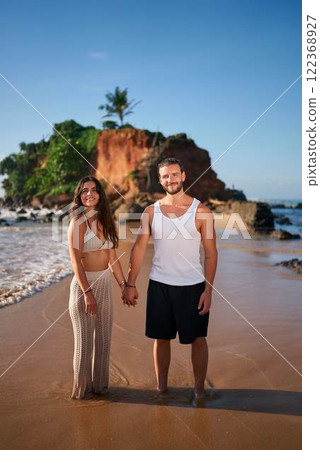 Couple holding hands on sandy beach, smiles facing camera. Man in tank, woman in dress enjoy romantic walk by sea. Sunset lights cliff backdrop. Honeymooners by ocean, travel love concept. Couple holding hands on sandy beach, smiles facing camera. Man in tank, woman in dress enjoy romantic walk by sea. Sunset lights cliff backdrop. Honeymooners by ocean, travel love concept. 122368927