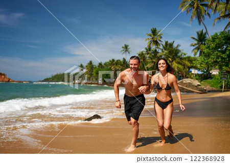 Fit couple running on tropical beach near ocean. Shirtless man and bikini-clad woman exercise by sea. Jogging duo enjoy coastline fitness. Active lifestyle, wellness by palm trees in sunshine. Fit couple running on tropical beach near ocean. Shirtless man and bikini-clad woman exercise by sea. Jogging duo enjoy coastline fitness. Active lifestyle, wellness by palm trees in sunshine. 122368928