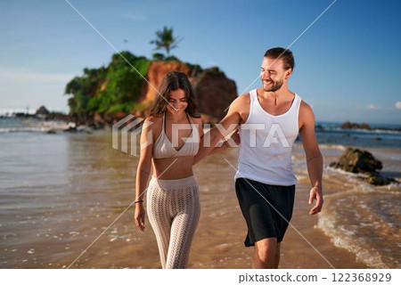 Fit couple hold hands, walk on tropical beach. Smiling woman in bikini, man in tank top enjoy seaside. Freelancers unwind on sandy shore, balance work, travel lifestyle. Remote job, leisure together. Fit couple hold hands, walk on tropical beach. Smiling woman in bikini, man in tank top enjoy seaside. Freelancers unwind on sandy shore, balance work, travel lifestyle. Remote job, leisure together. 122368929