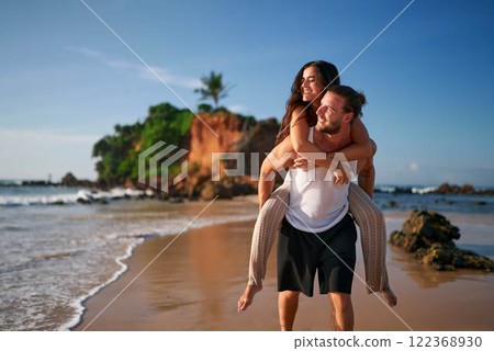 Romantic couple enjoys honeymoon on tropical beach. Man gives piggyback ride to smiling woman at sunset. Newlyweds walk by sea, love moments on exotic island. Happy bride, groom in casual wear. 122368930