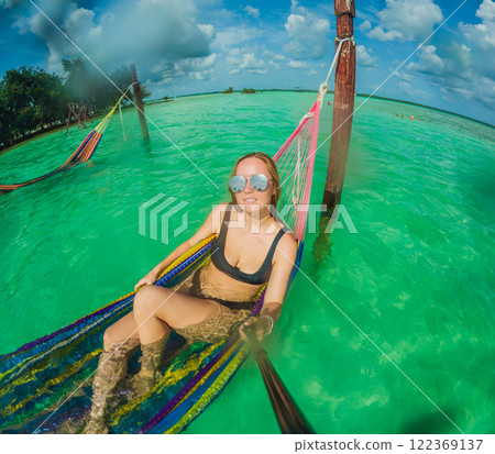 Woman tourist relaxing on a hammock above the turquoise waters of Lake Bacalar, Mexico. Peaceful retreat, tropical paradise, and relaxation concept in a serene natural setting Woman tourist relaxing on a hammock above the turquoise waters of Lake Bacalar, Mexico. Peaceful retreat, tropical paradise, and relaxation concept in a serene natural setting 122369137