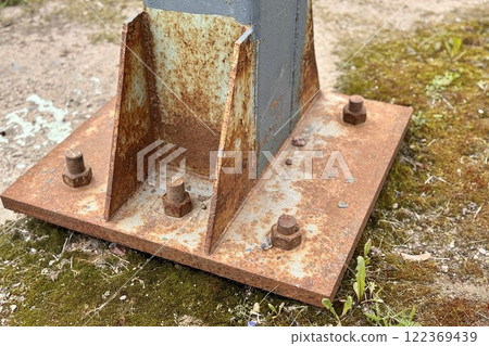 Metal pillar fastened to basement with anchor bolts and nuts. The rusty surface of the post and the fastenings are in places. The support of the iron black pillar on a green lawn in the park 122369439