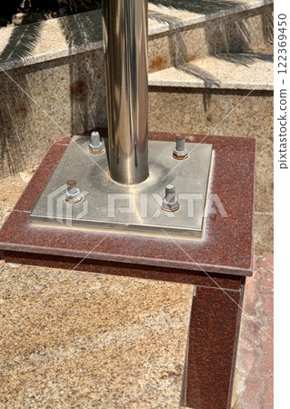Metal pillar fastened to basement with different anchor bolts and nuts. The support of the iron black pillar on a green lawn in city center Metal pillar fastened to basement with different anchor bolts and nuts. The support of the iron black pillar on a green lawn in city center 122369450