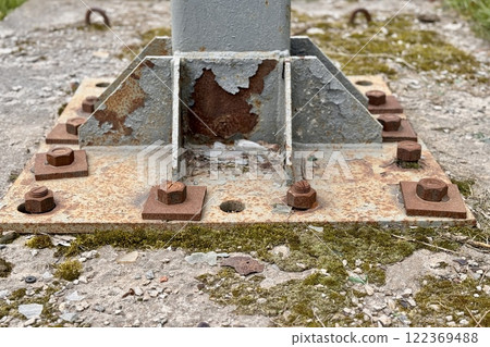 Metal pillar fastened to basement with anchor bolts and nuts. The rusty surface of the post and the fastenings are in places. The support of the iron pillar on a green lawn in the park. Close-up 122369488