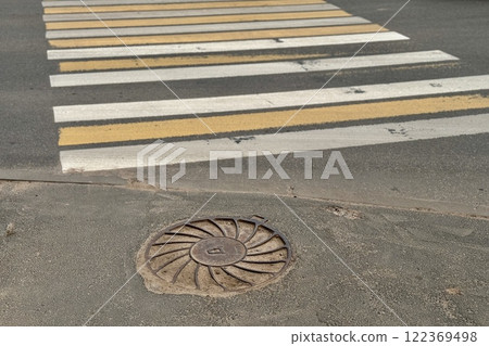 A sunny day view of a crosswalk with alternating yellow and white stripes beside a manhole cover 122369498