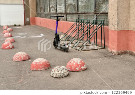 Red restrictive hemispheres on the asphalt road in the parking area of the car and the pedestrian area of walking people at the street. There is an unknown parked black scooter in front of them 122369499