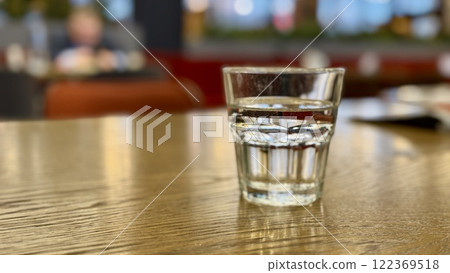 A clear glass glass with clean filtered water on a brown table in a restaurant against the background of a seated businessman. Healthy eating, strengthening the body and thirst for water. Close-up A clear glass glass with clean filtered water on a brown table in a restaurant against the background of a seated businessman. Healthy eating, strengthening the body and thirst for water. Close-up 122369518