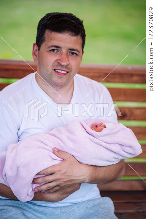 Smiling dad cradling newborn daughter in pink blanket, resting on wooden bench outdoors 122370829