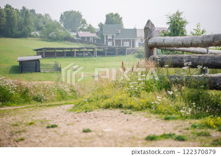Wooden fence flanking gravel path winding through peaceful russian village, revealing quaint rural homes amid pastoral landscape Wooden fence flanking gravel path winding through peaceful russian village, revealing quaint rural homes amid pastoral landscape 122370879