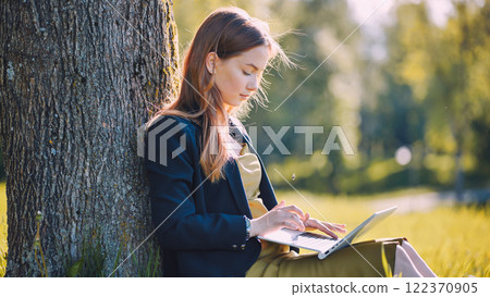 Young businesswoman using laptop while sitting under tree in park, enjoying fresh air and natural environment during work break 122370905
