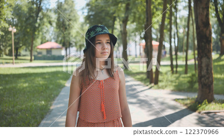Teen in bucket hat and orange dress strolling through verdant park, basking in golden sunlight and summer vibrancy Teen in bucket hat and orange dress strolling through verdant park, basking in golden sunlight and summer vibrancy 122370966