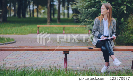 Young woman sits on a park bench, engrossed in her book, enjoying the peaceful atmosphere of nature 122370987