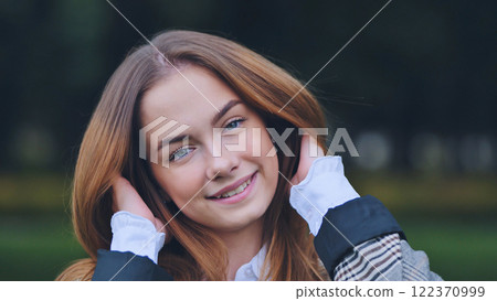Cheerful schoolgirl grasping long red hair, beaming with joy during bright, lush park outing under warm sunlight 122370999