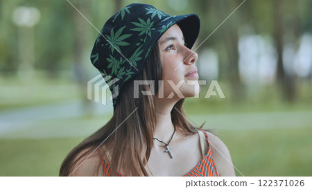 Teenage girl wearing cannabis patterned bucket hat, gazing upward pensively amid lush summer greenery Teenage girl wearing cannabis patterned bucket hat, gazing upward pensively amid lush summer greenery 122371026