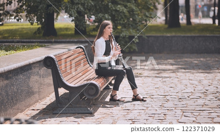 Young musician performing clarinet on wooden park bench, sunlight illuminating melodic moment of peaceful outdoor practice 122371029