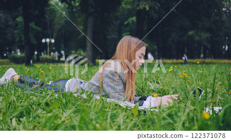 College student typing on laptop while relaxing on lush grass, embracing outdoor workspace with natural surroundings College student typing on laptop while relaxing on lush grass, embracing outdoor workspace with natural surroundings 122371078