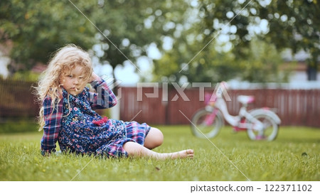 Blonde girl sitting on grass in backyard, touching her hair, with bicycle in background, enjoying a peaceful family day 122371102