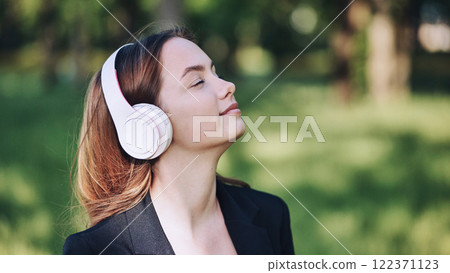 Young businesswoman wearing headphones is enjoying music or podcast in a green park, breathing fresh air Young businesswoman wearing headphones is enjoying music or podcast in a green park, breathing fresh air 122371123