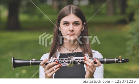 Young musician proudly holding her clarinet and posing in a vibrant green park under the warm sun on a beautiful summer day 122371229