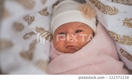 Newborn baby wearing a white hat with a small flower decoration, wrapped in a pink blanket, lying in a crib with feather patterns 122371238