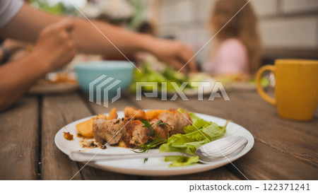 Rustic wooden table displaying hearty leftovers, grilled sausages, roasted potatoes, green salad on white plate, blurred family dining scene behind Rustic wooden table displaying hearty leftovers, grilled sausages, roasted potatoes, green salad on white plate, blurred family dining scene behind 122371241