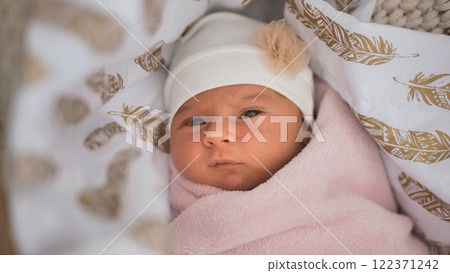 Sleeping newborn girl wearing white floral cap, nestled in pink blanket on delicate feather print bedding 122371242