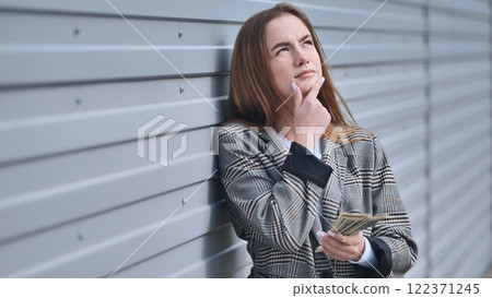 Serious businesswoman contemplating financial strategy, clutching dollar bills near weathered metal wall 122371245