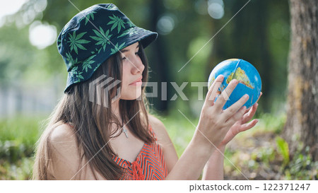 Young female contemplating miniature world globe, cannabis leaf beanie highlighting global awareness amid green park setting 122371247