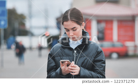 Young woman deeply focused on smartphone screen, navigating busy urban sidewalk with digital connectivity 122371270