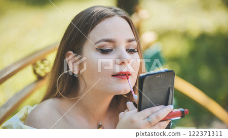 Young woman applying bright lipstick, checking reflection near sunlit urban street 122371311