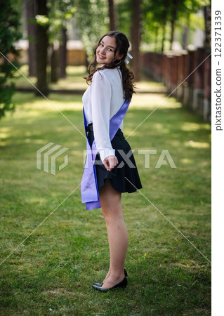 Smiling student in a purple sash rejoices on her final day of school, savoring the moment in a park 122371339