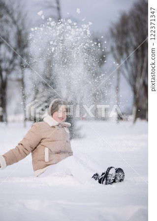 Happy schoolgirl throwing snow up into the air while sitting on a snow covered ground, enjoying winter holidays in a snowy park Happy schoolgirl throwing snow up into the air while sitting on a snow covered ground, enjoying winter holidays in a snowy park 122371347