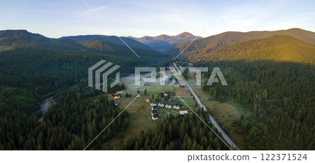 Aerial view of amazing scenery with small village houses between foggy dark mountain peaks covered with forest pine trees at autumn sunrise. Beautiful wild woodland with shining rays of light at dawn Aerial view of amazing scenery with small village houses between foggy dark mountain peaks covered with forest pine trees at autumn sunrise. Beautiful wild woodland with shining rays of light at dawn 122371524