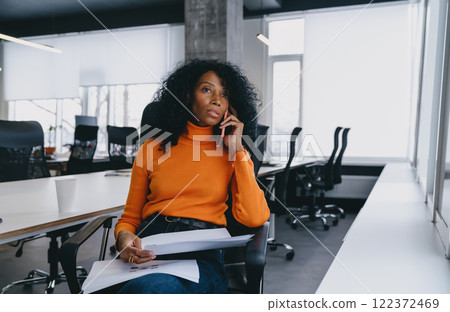 A pensive legal advisor in a vibrant orange sweater evaluates documents during a phone consultation in a contemporary office, hinting at discussions on car insurance or environmental policies. 122372469