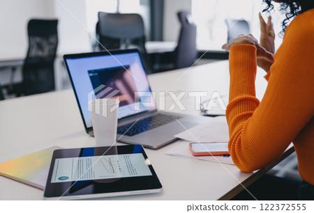 African American entrepreneur analyzing market trends on tablet at co-working, with laptop and smartphone, in orange sweater, embodying essence of private enterprise and agile business management. 122372555
