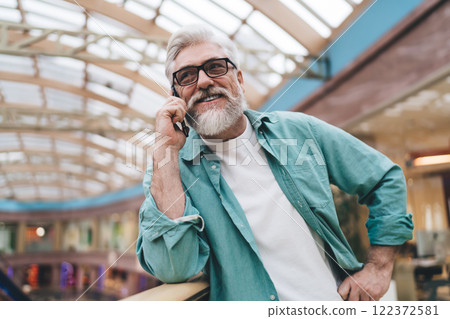 Mature Caucasian man in his 60s, with a well-groomed beard and glasses, speaks on a smartphone in vibrant co-working space, exemplifying active professional life and connectivity in modern retirement 122372581