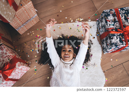 Happy cute black girl playing with stretched hands by colorful confetti on floor with Christmas gift boxes 122372597