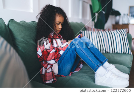 Cute black girl sitting on sofa while browsing smartphone 122372600