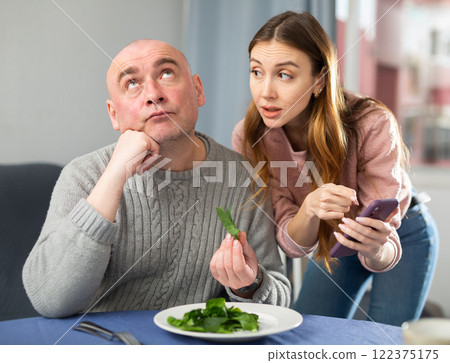 Wife with smartphone explaining health benefits of salad to her husband Wife with smartphone explaining health benefits of salad to her husband 122375175