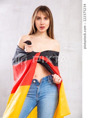 Flirting young woman in jeans with state flag of Germany against gray wall indoors 122375234